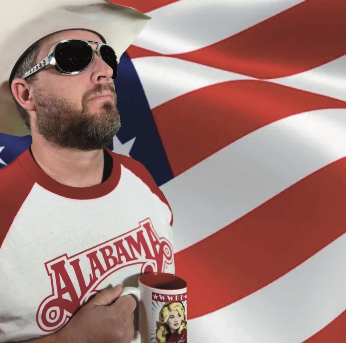Hoss Fly, a bearded man in a white cowboy hat and sunglasses, raising a mug in front of a large American flag